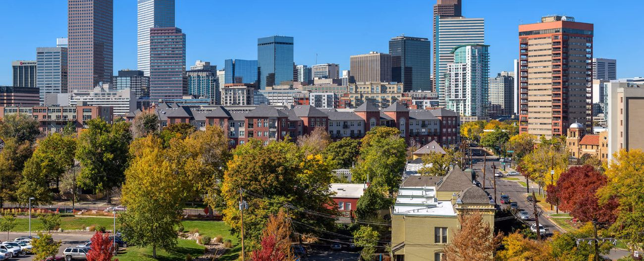 Aerial view of a downtown city skyline against a blue sky with suburban buildings mixed in with green trees next to a city street in the foreground